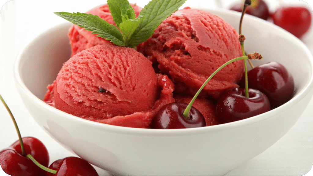 A bowl of vibrant red cherry sorbet garnished with fresh cherries and a mint sprig, set on a white surface.