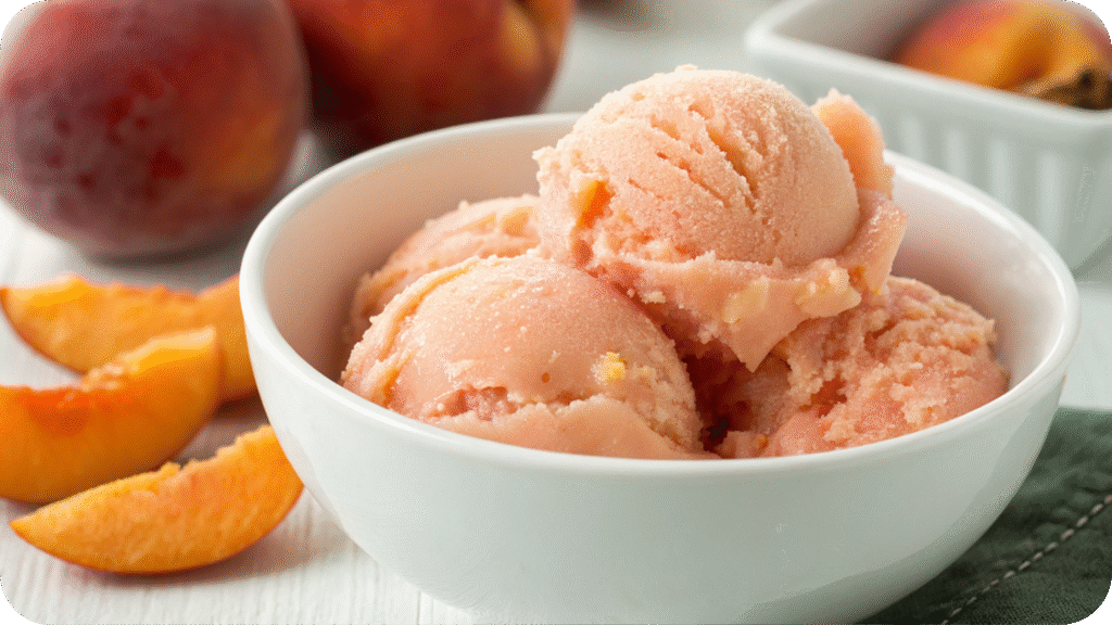 Creamy peach-colored sorbet scoops in a white bowl, surrounded by fresh peach slices and whole peaches.